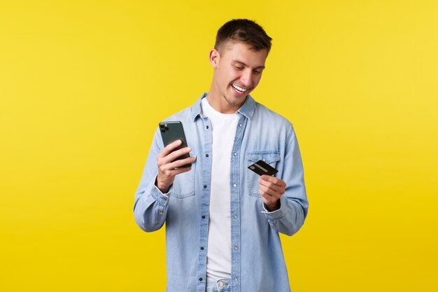 Concepto de tecnología, estilo de vida y publicidad. Hombre feliz guapo haciendo pedidos en línea, reservando boletos de avión con la aplicación de teléfono inteligente, mirando la tarjeta de crédito y sosteniendo el teléfono móvil, fondo amarillo.