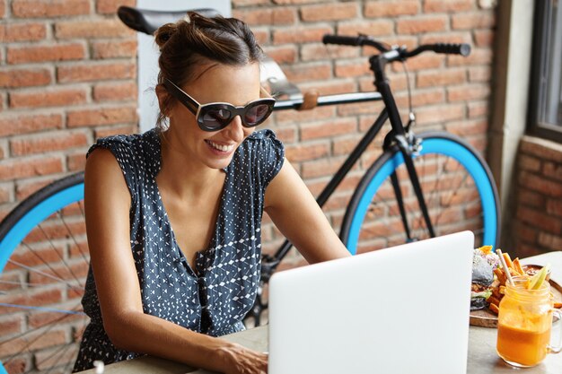 Concepto de tecnología y ocio. Mujer atractiva con elegantes gafas de sol haciendo videollamada a un amigo mientras almorzaba en la cafetería moderna