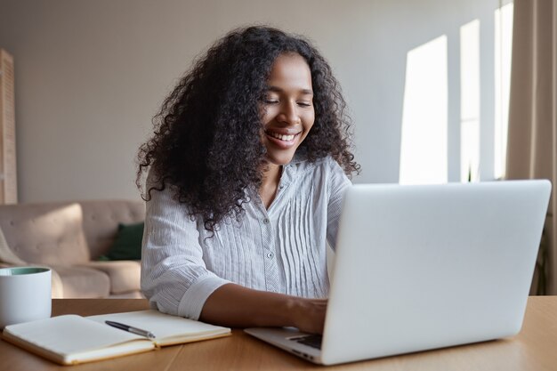 Concepto de tecnología, ocupación, autónomo y redes. Encantadora joven experta en marketing de piel oscura que trabaja a distancia de la oficina en casa, escribiendo en la computadora portátil, tomando notas y tomando café