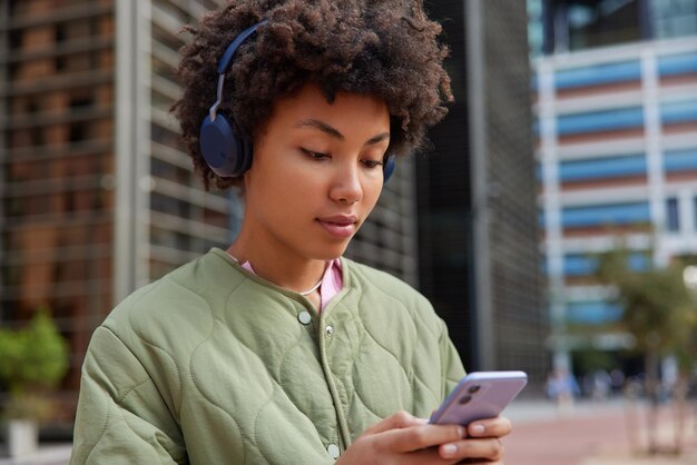 Concepto de tecnología y pasatiempo de estilo de vida de personas. Mujer de cabello rizado concentrada en la pantalla del celular escucha música a través de auriculares vestida con chaqueta y camina afuera contra los edificios de la ciudad.