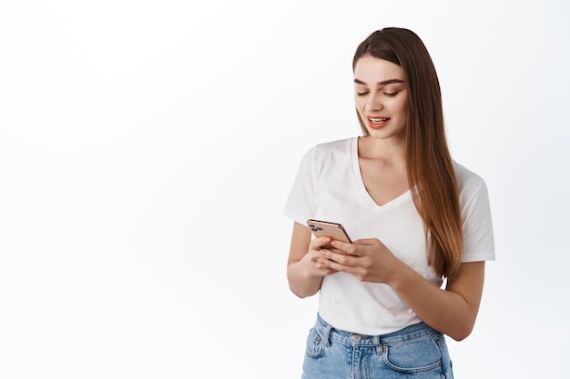 Concepto de tecnología y personas. Chica moderna con smartphone, mensajes de texto, lectura de mensajes en la pantalla del teléfono con cara relajada casual, de pie en camiseta contra el fondo blanco.