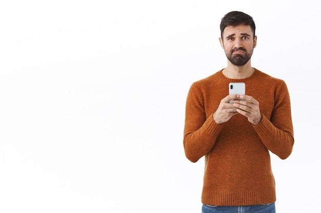 Concepto de tecnología, personas y comunicación. Retrato de un tipo triste y desafortunado con barba, haciendo muecas y frunciendo el ceño triste, recibe un mensaje perturbador, sosteniendo el teléfono móvil infeliz