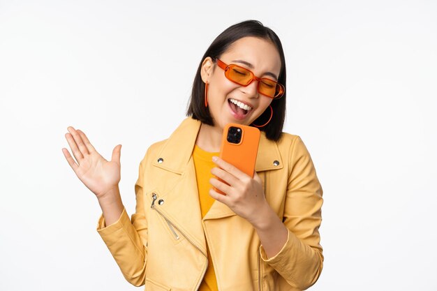 Concepto de tecnología y personas Feliz bailarina asiática con gafas de sol usando un teléfono inteligente cantando y sonriendo de pie sobre fondo blanco