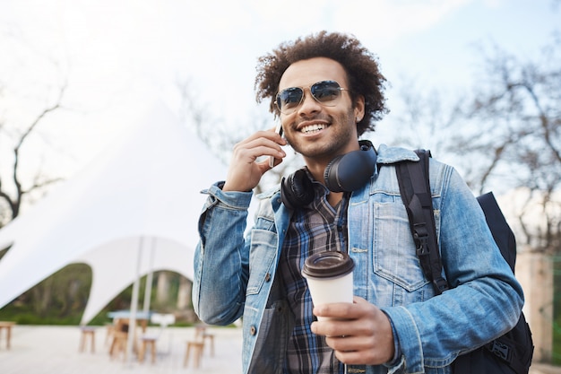 Concepto de tecnología y personas. Hombre joven guapo de piel oscura con cerdas y corte de pelo afro hablando por teléfono celular mientras toma café y camina por la ciudad, con mochila y abrigo de mezclilla.