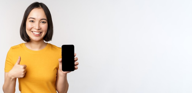 Concepto de tecnología y personas Mujer joven sonriente que muestra el pulgar hacia arriba y la interfaz de la aplicación de teléfono móvil de la pantalla del teléfono inteligente de pie contra el fondo blanco