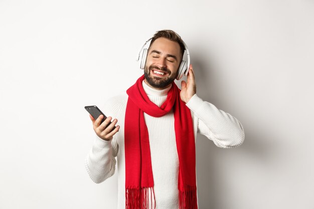 Concepto de tecnología y vacaciones de invierno. Hombre satisfecho escuchando música en auriculares con los ojos cerrados, sonriendo con placer, sosteniendo smartphone, fondo blanco.