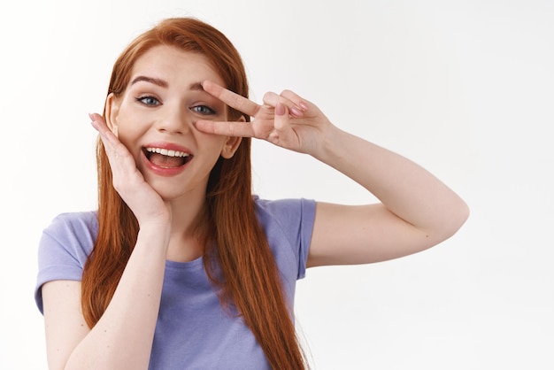 Concepto de ternura y maquillaje de belleza Mujer pelirroja caucásica de buen aspecto alegre con camiseta morada que toca la piel pura y limpia sin imperfecciones muestra un gesto de paz sobre los ojos sonriendo feliz