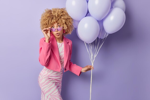 Concepto de tiempo de fiesta Una mujer joven de cabello rizado asombrada que usa un vestido tiene una figura perfecta que mira fijamente sorprendentemente mantiene la mano en las gafas de sol sostiene un montón de globos inflados listos para la celebración del cumpleaños