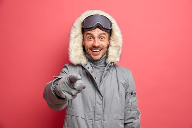 Concepto de tiempo de invierno feliz. El hombre europeo alegre en ropa de abrigo indica directamente con expresión alegre que ve algo muy agradable en el frente.