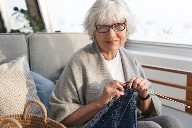 Concepto de tiempo libre, pasatiempo, relajación, edad y artesanía. Alegre encantadora mujer de mediana edad jubilada relajándose en casa, tejiendo un suéter cálido para la venta, vistiendo elegantes gafas y sonriendo