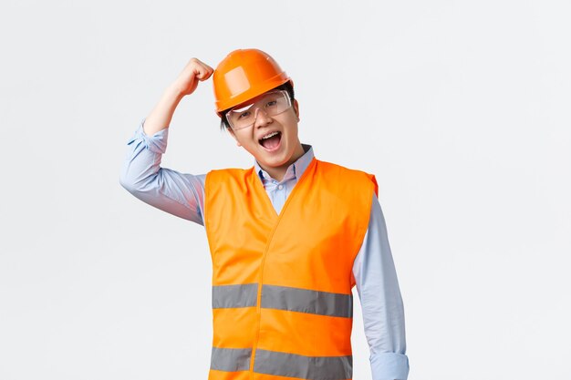 Concepto de trabajadores industriales y del sector de la construcción. Ingeniero masculino entusiasta en ropa reflectante golpeando el casco y sonriendo, asegúrese de que los empleados trabajen con buena protección, fondo blanco.