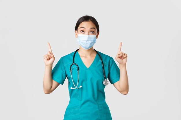 Concepto de trabajadores de la salud de la enfermedad del coronavirus covid19 Intrigada y emocionada mujer asiática doctora enfermera con máscara médica y guantes mirando y señalando con el dedo hacia el fondo blanco