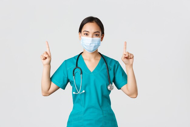 Concepto de trabajadores de la salud de la enfermedad del coronavirus covid19 Joven enfermera médica asiática profesional con máscara médica y batas señalando con el dedo hacia arriba mostrando publicidad
