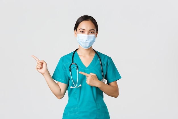 Concepto de trabajadores de la salud de la enfermedad del coronavirus covid19 Profesional médica asiática segura de sí misma doctora con máscara médica y batas señalando con el dedo a la izquierda mostrando publicidad