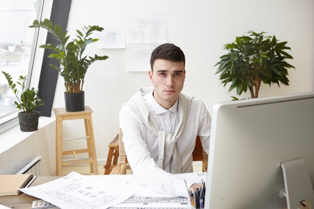 Concepto de trabajo, carrera y éxito. Filmación en interiores del talentoso joven arquitecto con camisa blanca y un suéter atado alrededor de su cuello usando un programa CAD en la computadora mientras trabajaba en un proyecto de construcción