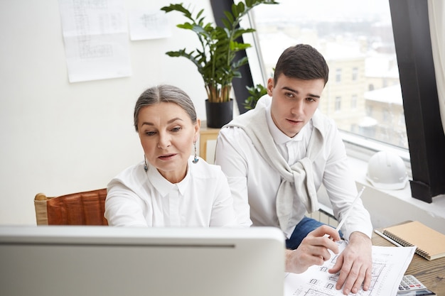 Concepto de trabajo en equipo, cooperación y colaboración. Equipo de dos arquitectos expertos creativos, mujer senior y hombre joven sentados frente a la computadora, intercambiando ideas y discutiendo dibujos juntos