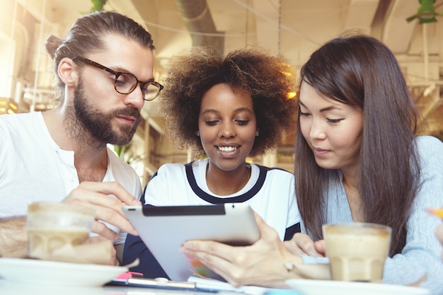 Concepto de trabajo en equipo y cooperación. Tres talentosos jóvenes ambiciosos intercambiando ideas, teniendo una discusión sobre un proyecto común usando una tableta digital en la cafetería.