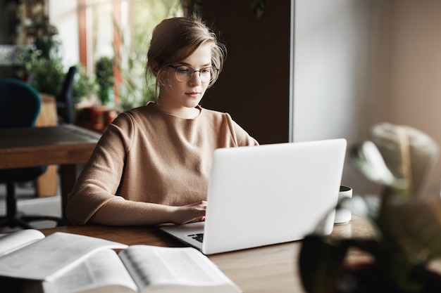 Concepto de trabajo, estilo de vida y negocios. Atractiva mujer europea enfocada en gafas de moda sentado en un café cerca de la computadora portátil, trabajando en el cuaderno, rodeado de libros, tomando notas.