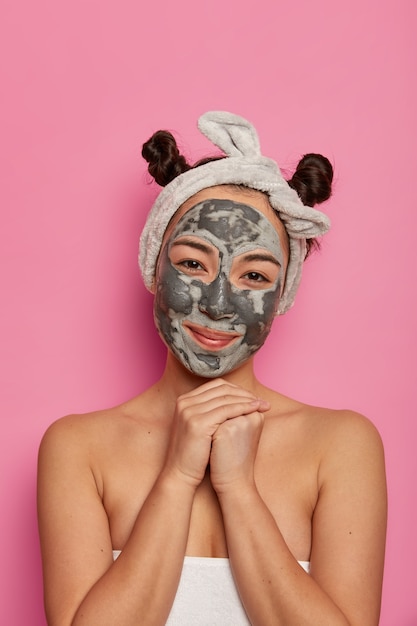 Concepto de tratamiento de personas, cuidado de la piel y spa. La modelo femenina satisfecha mantiene las manos debajo de la barbilla, aplica mascarilla para una piel pura perfecta y reduce las arrugas, se mantiene fresca y joven, mantiene una higiene saludable