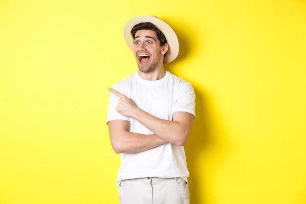 Concepto de turismo y estilo de vida. Chico guapo emocionado con sombrero de paja mirando publicidad, apuntando y mirando el logo de la esquina superior izquierda, fondo amarillo.