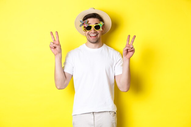 Concepto de turismo y estilo de vida. Hombre feliz disfrutando de viaje, con sombrero de verano y gafas de sol, posando con signos de paz para la foto, fondo amarillo