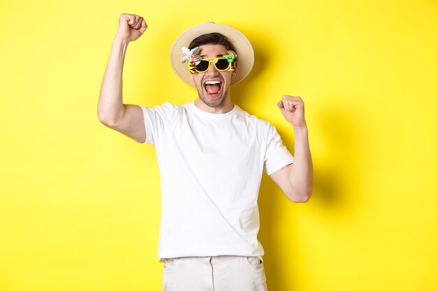 Concepto de turismo y estilo de vida. Hombre feliz ganando el viaje al resort, gritando que sí y levantando las manos, triunfando, con gafas de sol y sombrero de verano, fondo amarillo.