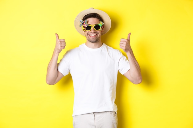 Concepto de turismo y estilo de vida. Imagen de un turista sonriente mostrando el pulgar hacia arriba, disfrutando del viaje y recomendando, con sombrero de verano y gafas de sol, fondo amarillo.