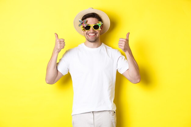 Concepto de turismo y estilo de vida. Imagen de un turista sonriente mostrando el pulgar hacia arriba, disfrutando del viaje y recomendando, con sombrero de verano y gafas de sol, fondo amarillo.