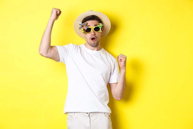 Concepto de turismo y estilo de vida. Turista de chico feliz disfrutando del viaje, alentándote, puñetazo y triunfando, yendo de viaje con sombrero de verano y gafas de sol, fondo amarillo.