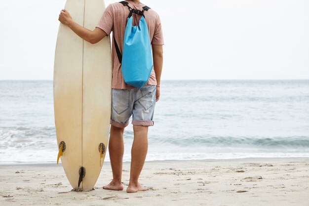 Concepto de turismo, ocio y estilo de vida saludable. Vista posterior del joven surfista de pie descalzo en la orilla arenosa, frente al vasto océano y sosteniendo su tabla de surf