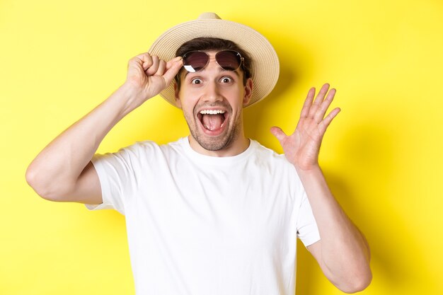 Concepto de turismo y vacaciones. Gafas de sol de despegue turístico hombre emocionado y mirando sorprendido por la oferta promocional, de pie sobre fondo amarillo.