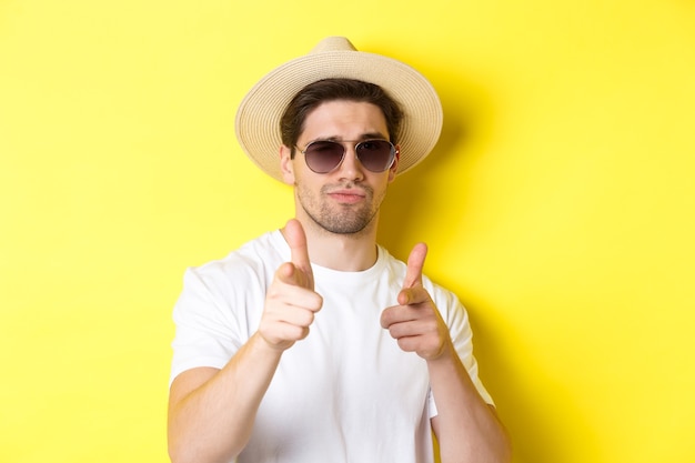 Concepto de turismo y vacaciones. Primer plano de chico cool con sombrero de verano y gafas de sol apuntando con armas de dedo a la cámara, de pie sobre fondo amarillo.