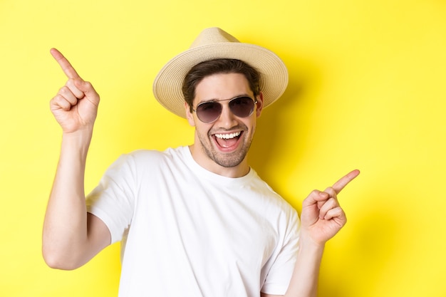 Concepto de turismo y vacaciones. Primer plano del hombre disfrutando de las vacaciones de viaje, bailando y señalando con el dedo hacia los lados, con gafas de sol con sombrero de paja.