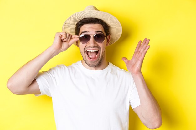 Concepto de turismo y vacaciones. Primer plano de hombre sorprendido gritando de alegría, disfrutando de las vacaciones, con gafas de sol con sombrero de verano, fondo amarillo.