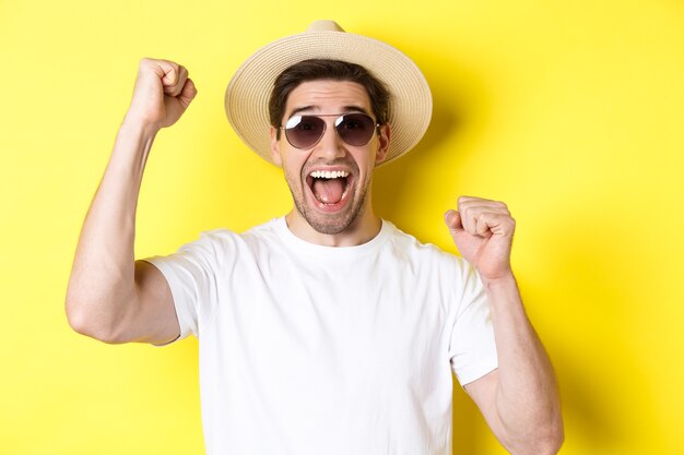 Concepto de turismo y vacaciones. Turista masculino feliz celebrando sus vacaciones, levantando las manos y gritando de alegría, con gafas de sol con sombrero de paja.