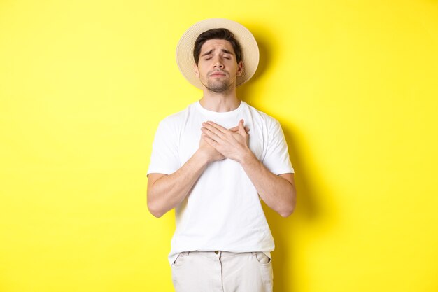 Concepto de turismo y verano. Hombre romántico con sombrero de paja mirando nostálgico, cerrar los ojos y cogidos de la mano en el corazón, de pie contra el fondo amarillo.