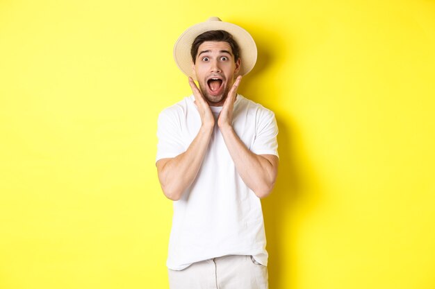 Concepto de turismo y verano. Modelo masculino sorprendido con sombrero de paja, mirando asombrado por la oferta especial, de pie contra el fondo amarillo