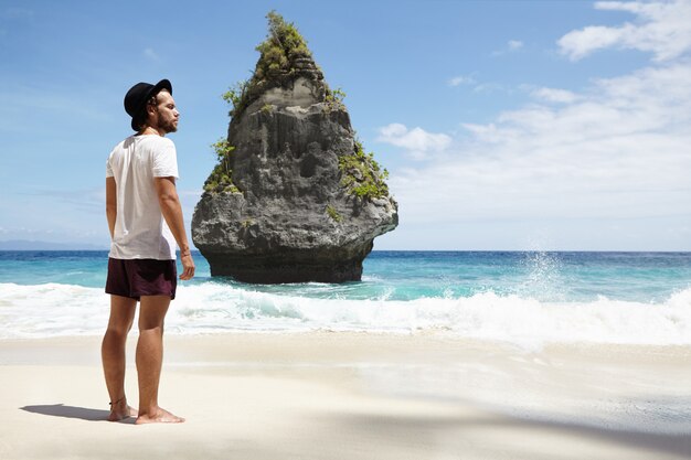 Concepto de turismo, viajes y vacaciones. Joven modelo masculino caucásico con sombrero negro y ropa casual posando descalzo sobre la arena mojada con una isla rocosa frente a él mientras grandes olas golpean la orilla