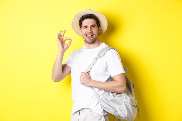 Concepto de turismo, viajes y vacaciones. Turista feliz yendo de vacaciones, sosteniendo la mochila y mostrando un signo bien sonriendo satisfecho, de pie contra el fondo amarillo.