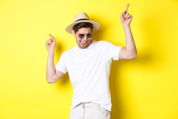 Concepto de turismo, viajes y vacaciones. Turista masculino disfrutando de las vacaciones, bailando con sombrero de paja y gafas de sol, señalando con el dedo hacia los lados.