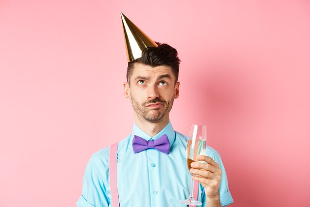 Concepto de vacaciones y celebración. Chico gruñón con sombrero de fiesta de cumpleaños y sosteniendo una copa de champán, mirando hacia arriba con cara escéptica, de pie sobre fondo rosa.