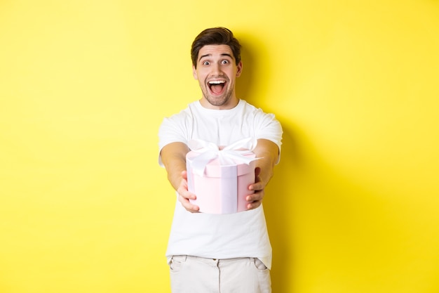 Concepto de vacaciones y celebración. Hombre feliz deseando feliz año nuevo, dándole un regalo, de pie sobre fondo amarillo.