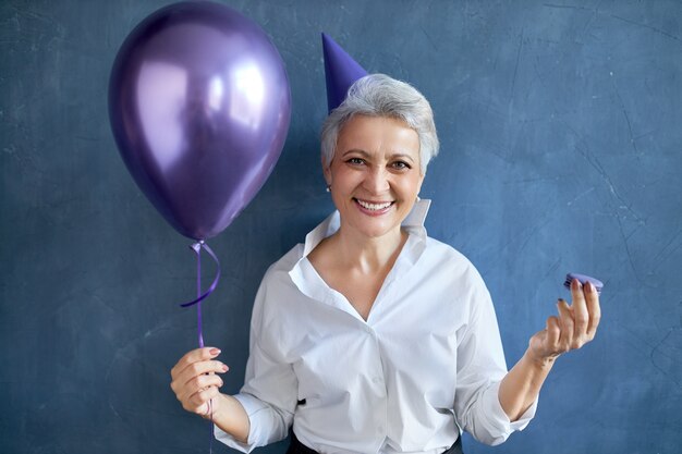 Concepto de vacaciones, celebraciones y ocasiones especiales. Retrato de mujer jubilada caucásica divertida emocional en ropa festiva sosteniendo globo de helio