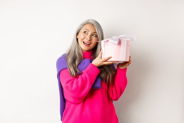 Concepto de vacaciones y día de San Valentín. Feliz mujer madura asiática agitando la caja con el regalo, adivinando qué dentro del presente, de pie sobre fondo blanco.