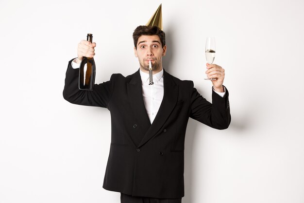 Concepto de vacaciones y estilo de vida. Chico guapo celebrando un cumpleaños, soplando el silbato de la fiesta y sosteniendo champán, diciendo un brindis, de pie en traje sobre fondo blanco.