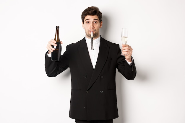 Foto gratuita concepto de vacaciones, fiesta y celebración. retrato de chico guapo en traje negro, levantando una botella de champán y vidrio, soplando un silbato de fiesta, teniendo un cumpleaños, de pie sobre fondo blanco.