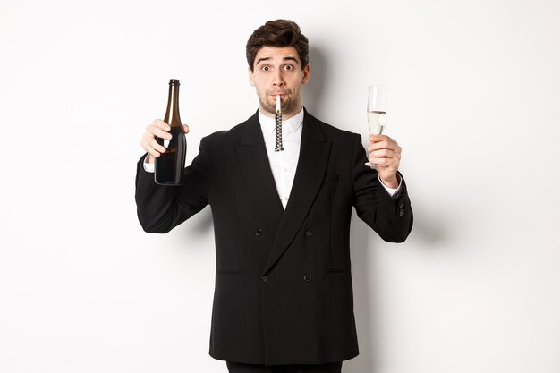 Concepto de vacaciones, fiesta y celebración. Retrato de chico guapo en traje negro, levantando una botella de champán y vidrio, soplando un silbato de fiesta, teniendo un cumpleaños, de pie sobre fondo blanco.