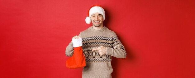 Concepto de vacaciones de invierno, año nuevo y celebración. Un apuesto hombre sonriente preparó regalos para los niños, apuntando a una bolsa de almacenamiento de Navidad, de pie sobre un fondo rojo.