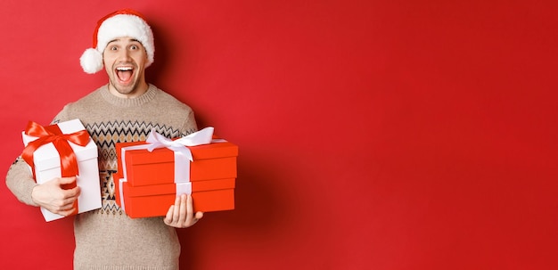 Foto gratuita concepto de vacaciones de invierno año nuevo y celebración imagen de hombre asombrado y feliz en suéter y sombrero de santa con regalos de navidad y gritando de alegría fondo rojo