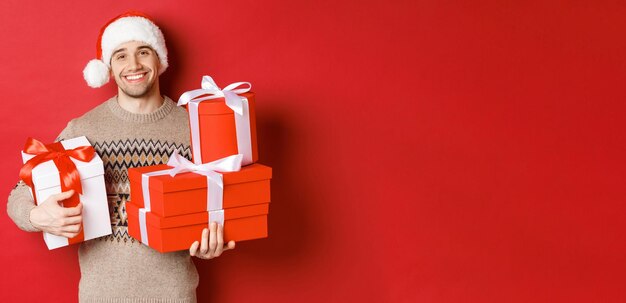 Concepto de vacaciones de invierno, año nuevo y celebración. Retrato de hombre sonriente encantador preparó regalos para Navidad, sosteniendo regalos y mirando a cámara conmovedor, fondo rojo.